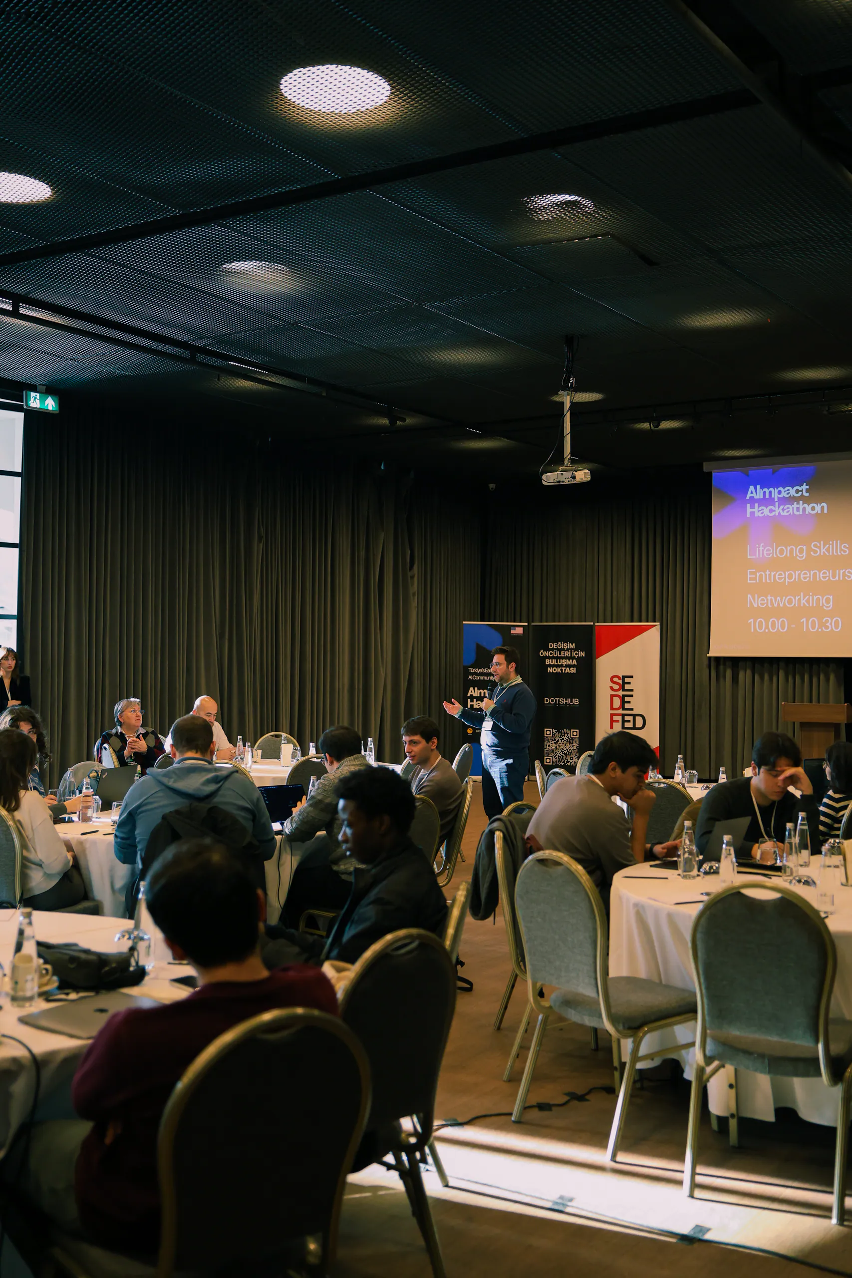 AImpact programında yuvarlak masalarda çalışan katılımcılar ve sahnedeki sunum ekranı